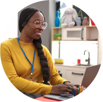 Female teacher seated at classroom table working on laptop, smiling